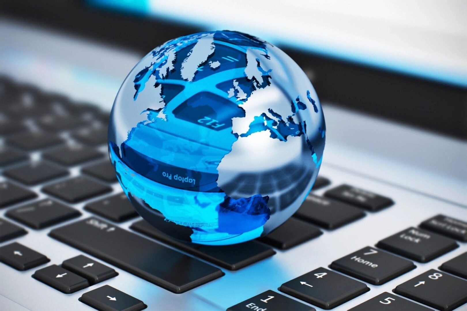 A small glass globe with blue continents sits on a computer keyboard, reflecting parts of the keyboard and screen in its surface.