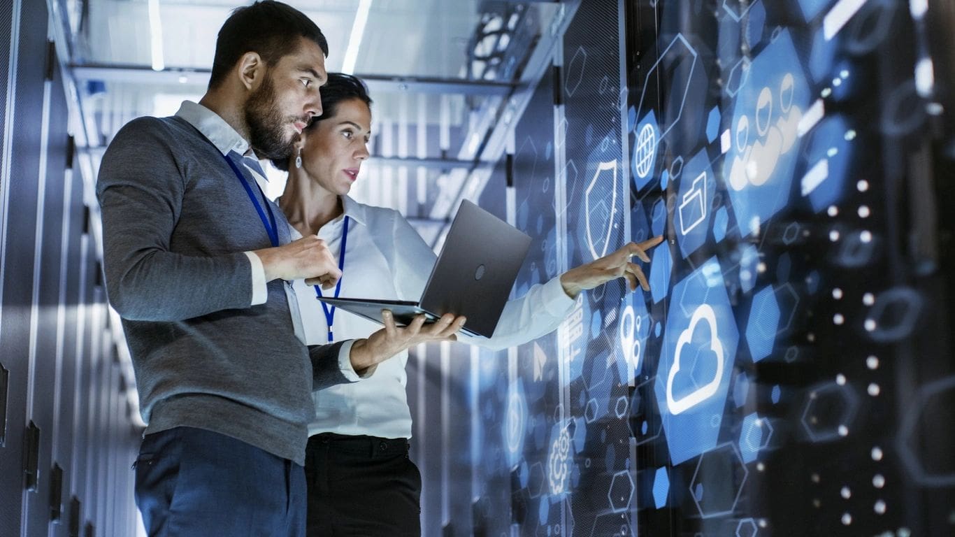 qtq80-T6ocuG Two professionals stand in a server room, looking at digital information on a large screen. One holds a laptop while the other points at cloud and data icons on the display wall.