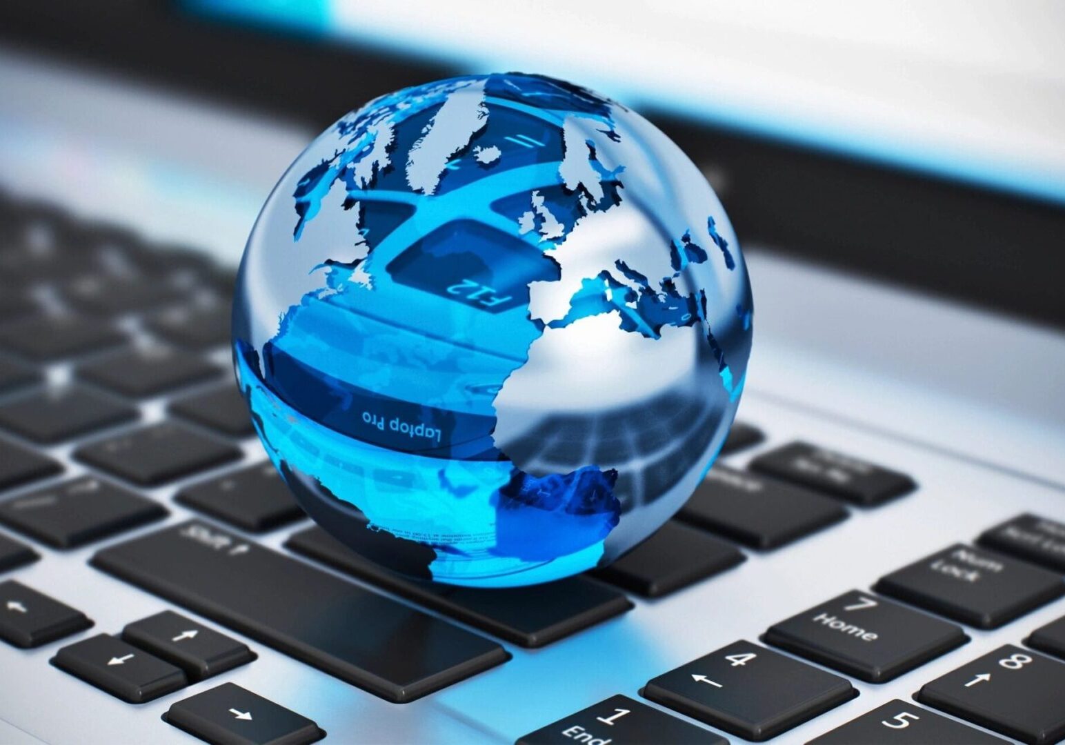 A small glass globe with blue continents sits on a computer keyboard, reflecting parts of the keyboard and screen in its surface.