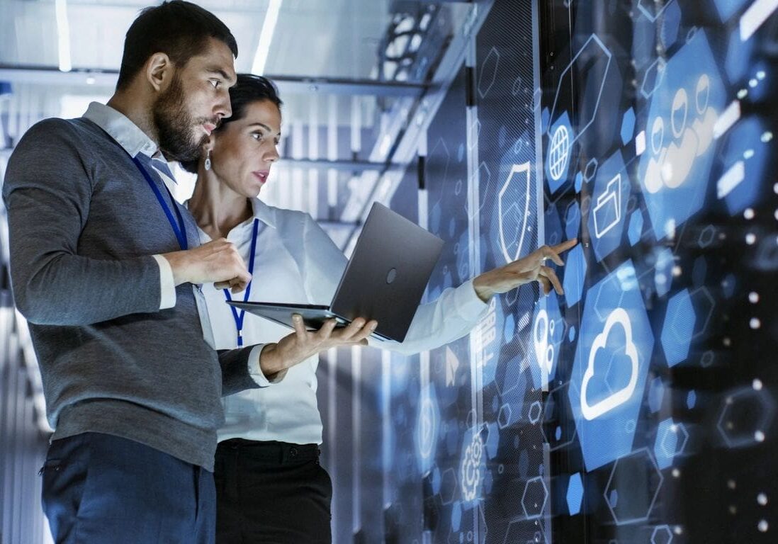Two professionals stand in a server room, looking at digital information on a large screen. One holds a laptop while the other points at cloud and data icons on the display wall.
